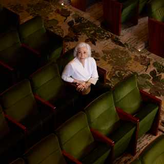 Fernanda Montenegro, with short grey hair and a white top, sits in a row of green theatre seats looking up at the camera.