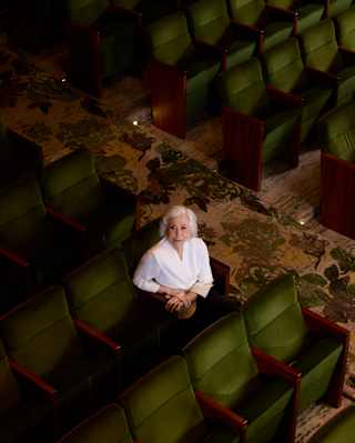 Fernanda Montenegro, with short grey hair and a white top, sits in a row of green theatre seats looking up at the camera.