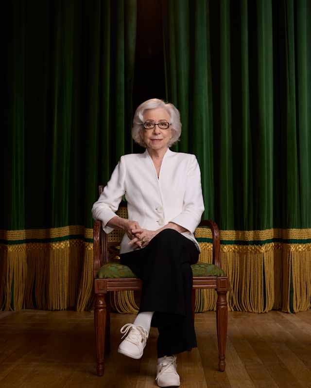 Fernanda Montenegro, with grey hair, glasses, dark trousers and white top, sits elegantly on a chair on stage, looking ahead.