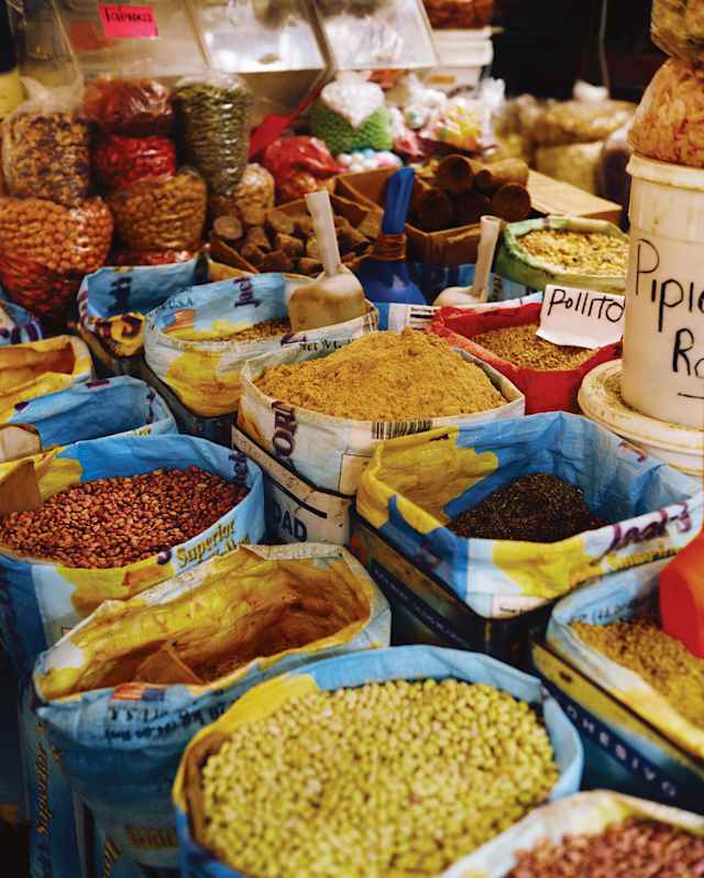 Bright image of two rows of open sacks containing grains and spices, and bags of dried beans, seen at a Mexican market stall.