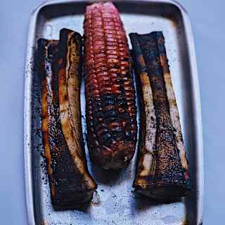 An cob of grilled red corn sits between two charred bones, with marrow exposed, on a silver cooking tray, seen from above.