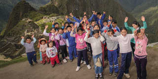 A large group of children of all ages cheer and smile to camera in front of Machu Picchu and the imposing Huayna Picchu peak.