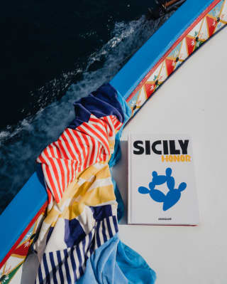 A copy of Sicily Honour rests on the deck of a boat with a pretty, hand-painted side, next to a red, blue and yellow towel.