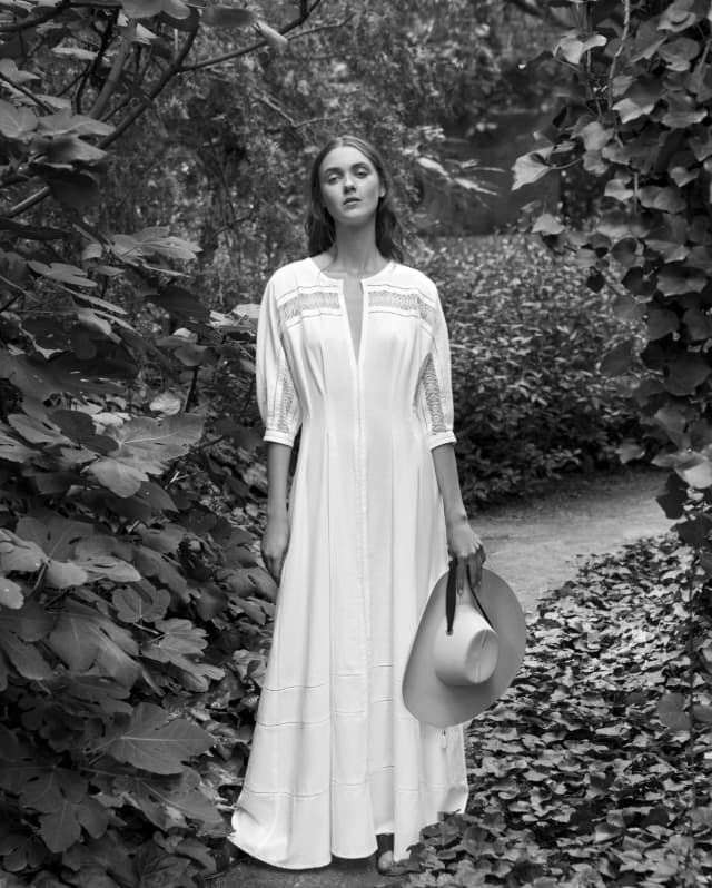 Pictured in black and white, a model wears a white cotton Concha dress with lace-like embroidery, by Collectiva Concepción.