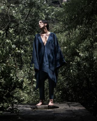 A barefoot female model poses in a black poncho with a deep v-neck by Collectiva Concepció, worn over black trousers.