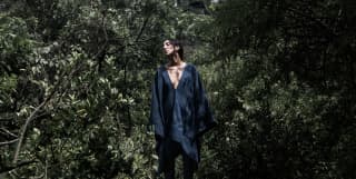 A barefoot female model poses in a black poncho with a deep v-neck by Collectiva Concepció, worn over black trousers.