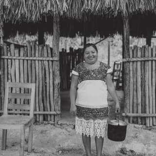 Black and white image of a Yucatan Peninsula mama, standing in front of a wood and thatch cabana holding a handled pot.