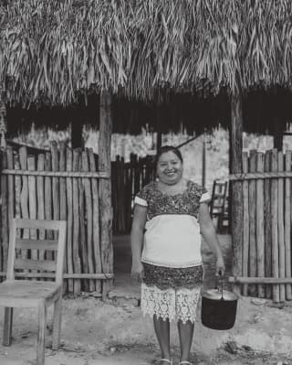 Black and white image of a Yucatan Peninsula mama, standing in front of a wood and thatch cabana holding a handled pot.