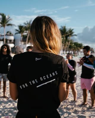A group of people wearing Parley For The Ocean shirts gather in a circle on the beach, viewed from behind a female member.