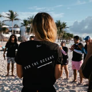 A group of people wearing Parley For The Ocean shirts gather in a circle on the beach, viewed from behind a female member.