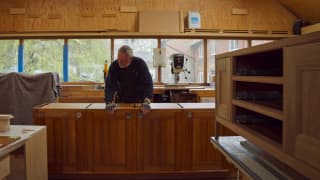 Furniture maker William Garvey, in a black jumper and fingerless gloves, stands behind a row of cabinets in his workshop.