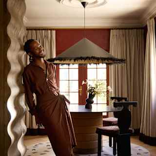 Wearing a brown casual suit, Thebe Magugu leans against a wavy wooden wall arch in the Mount Nelson Hotel suite he designed.