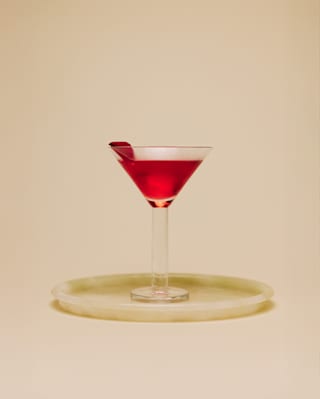 Eye-level view of a glowing, deep red Rose Martini cocktail, garnished with a petal, served in a thick-stemmed Martini glass.