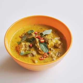 Close-up of Panang Curry with meat and slices of red chilli and curry leaves, served in a yellow bowl, seen from above.