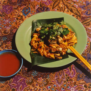 A crispy oyster omelette is served on a green plate, set on a Malay fabric cloth with chopsticks and dipping sauce.