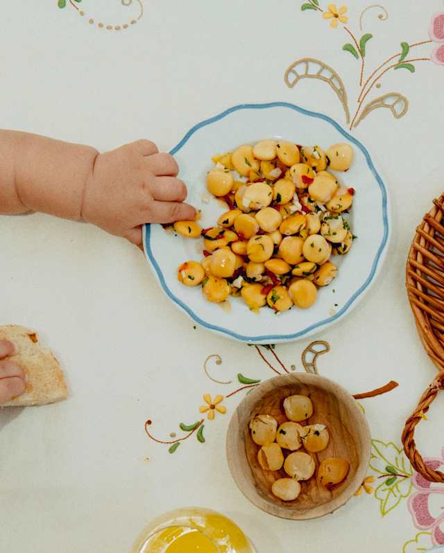A very young child's hand reaches for a bowl of cooked yellow lupini beans tossed in seasoning, seen from above.