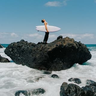 A man with a shortie surfboard under his arm stands, looking at the sky ,on a large basaltic rock surrounded by foaming sea.
