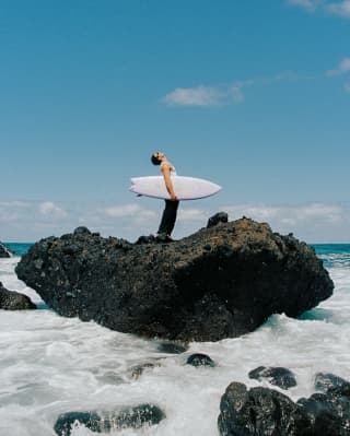 A man with a shortie surfboard under his arm stands, looking at the sky ,on a large basaltic rock surrounded by foaming sea.