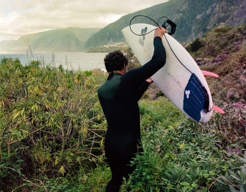 A man in a wetsuit carries his surfboard, with pink fins, through Madeira's undergrowth towards the ocean, seen from behind.