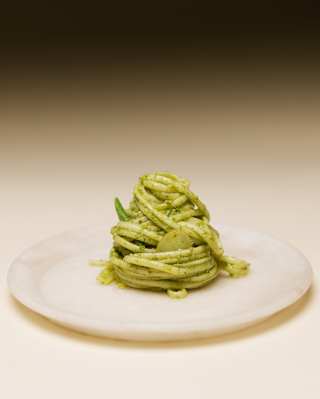 A perfect twirled mound of trenette pasta in a fresh, vibrant pesto sauce, served on a simple white plate, seen in close-up.