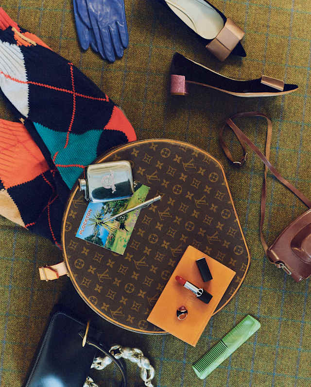 A Louis Vuitton round bag sits on a green textured surface, surrounded by a colourful jumper, gloves, shoes, a wallet, lipstick, sunglasses, keys, and a green comb. Items are arranged in a stylish overhead layout.