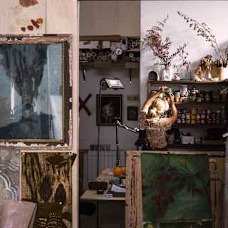 Elena Carozzi, with chignon-ed blond hair looks for pigments on an art shelf, seen from behind in her busy wallpaper studio.