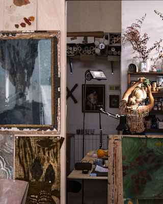 Elena Carozzi, with chignon-ed blond hair looks for pigments on an art shelf, seen from behind in her busy wallpaper studio.
