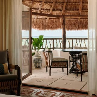 Looking through the doors of an Ocean View Junior Suite to a balcony with a straw canopy, seating and views onto the ocean.