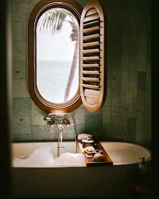 A shutter opens to reveal sea views through an oval window set in the wall above a bubbling bath with a cross-tub soap tray.