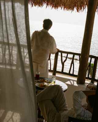 High angle view of a man in white top and trousers leaning on his balcony rail as he gazes at sun-bleached sea below.