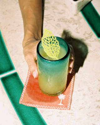 Close-up of a guest's hand holding a cocktail from Freddy's Bar, served in a thick, blue-tinted glass with a leaf decoration.