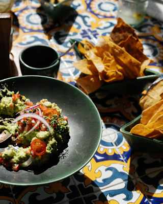 Tomato, onion and avocado salsa with two bowls of tortilla chips create a vibrant lunch on a yellow and blue-tiled table.