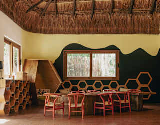 A view into the kids club with a table and eight red-framed chairs, wood-hut reading space and hexagonal wooden shelves.
