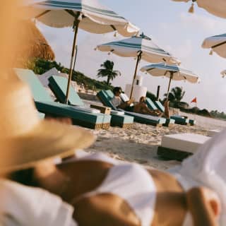 A couple in chairs relax in parasol shade with a soft-focus close-up on a woman lying on daybed with a sunhat over her face.