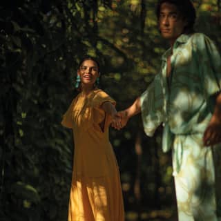 A dark-haired woman with turquoise earrings and a yellow dress leads a dark man in a soft green suit into the shady gardens.