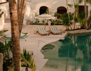 Two green and white striped sun loungers sit beneath a parasol by the green mirror-glass pool, flanked by towering palms.