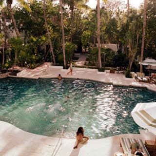In a pool scene shot from above, a man lies on a lounger, a woman sits at the edge and guests swim in the sun-dappled waters.