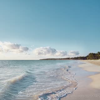 View along the waterline as a shallow wave rolls towards the white sand beach, with a blue sky and small, fluffy clouds.