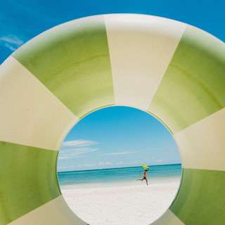A man runs across a white beach by an azure sea, holding a yellow kite in his hand, viewed through an inflatable rubber ring.