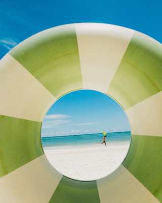 A man runs across a white beach by an azure sea, holding a yellow kite in his hand, viewed through an inflatable rubber ring.