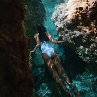 A woman in a white swimming costume swims between ancient rocks in a cenote with crystal blue water, dappled with sun glints.