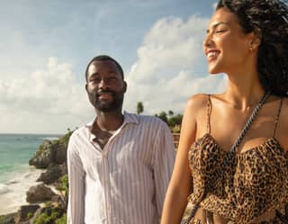 An a coastal path, a man in a soft candy-stripe shirt walks behind a woman in an animal-print dress who smiles at the view.