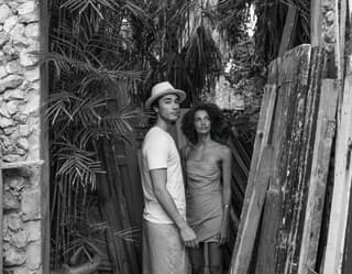 In black and white, a casually-dressed man and woman, standing in a cluttered garden yard, look back through a stone doorway.