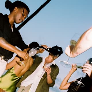Low angle, diagonal shot of a man in black pouring sparkling wine as guests raise a toast at their dining table on the beach.