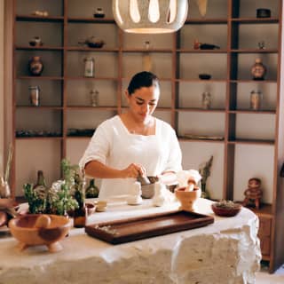 Wearing a white tunic, a female member of the spa staff grinds herbs on the Apothecary table under the glowing ceramic lamps.