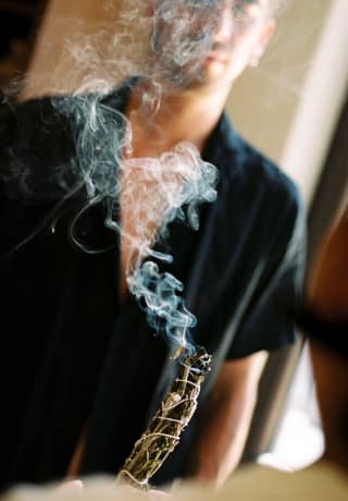 A man in a black shirt watches as a smudge stick of dried herbs and flowers burns with plumes of smoke at a welcome ceremony.>