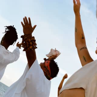Low angle view of the Shaman with a conch shell in his raised hands, flanked by guests with their arms held up as they chant.