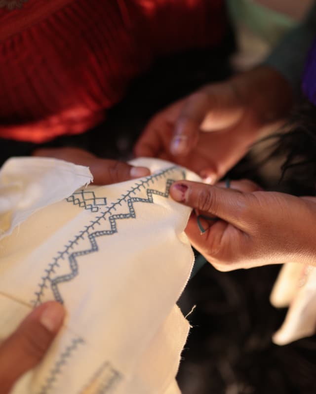 An embroiderer stitches track and zigzag patterns on to white cotton as another craftsperson holds it taut, seen in closeup.