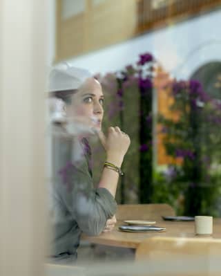 Chef Pía León looks pensive as she sits at a table, viewed from the outside through a window that reflects strips of sky.