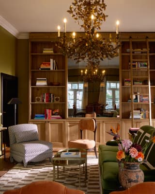 An oak-branch chandelier dominates an eclectic living room with book shelves, green sofa, orange chairs, flowers and art.